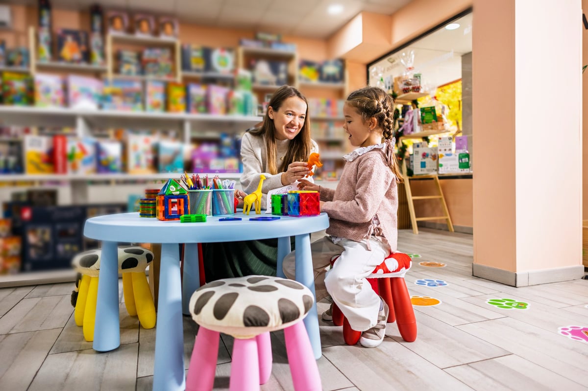 Mother and daughter happily engaging in creative play with magnetic building blocks and animal figures at a small table inside a colorful toy store or educational center