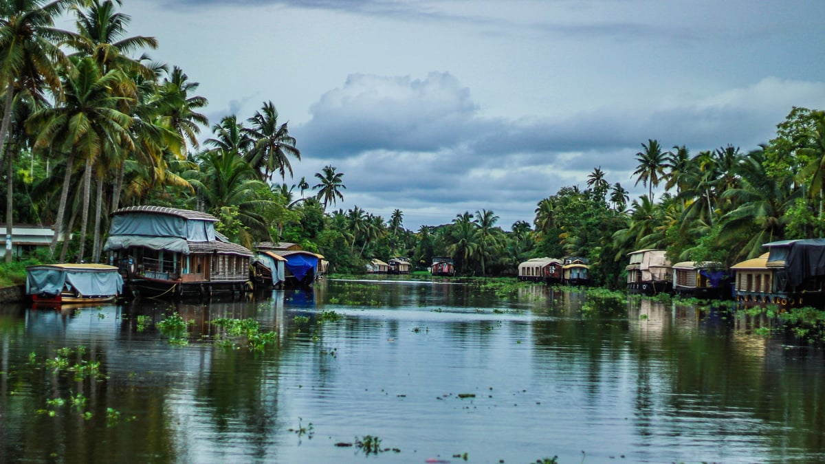Houseboats moored along the tranquil Kerala backwaters under dramatic monsoon clouds, surrounded by lush coconut palms and calm reflective waters in Alappuzha, Kerala, India