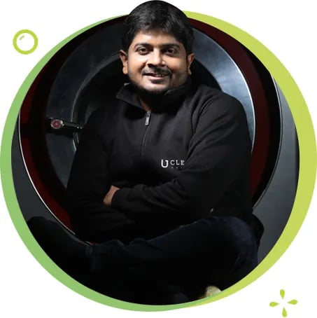 Man smiling at camera wearing black hoodie, seated against circular lime green frame backdrop