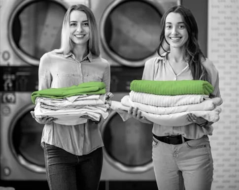 Two smiling women holding stacks of folded laundry in front of commercial washing machines at a laundromat