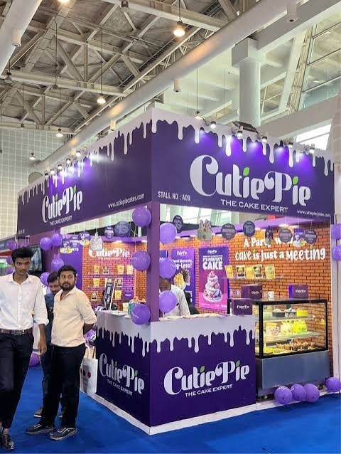 Cutie Pie bakery booth with purple frosting-drip design and white lettering at an indoor trade show or exhibition