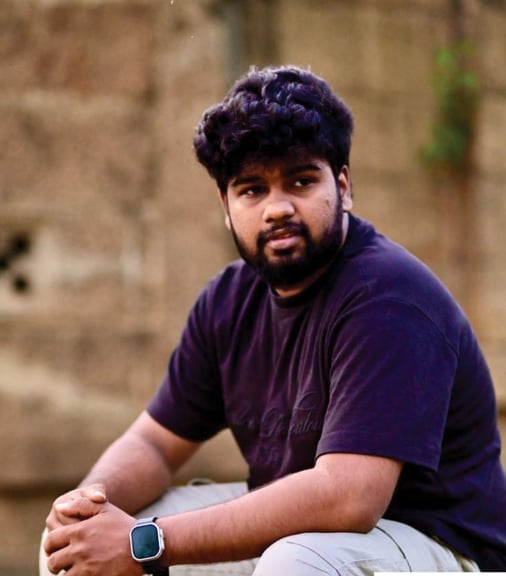 Man with dark curly hair and beard wearing purple t-shirt and smartwatch, posing against weathered brick wall